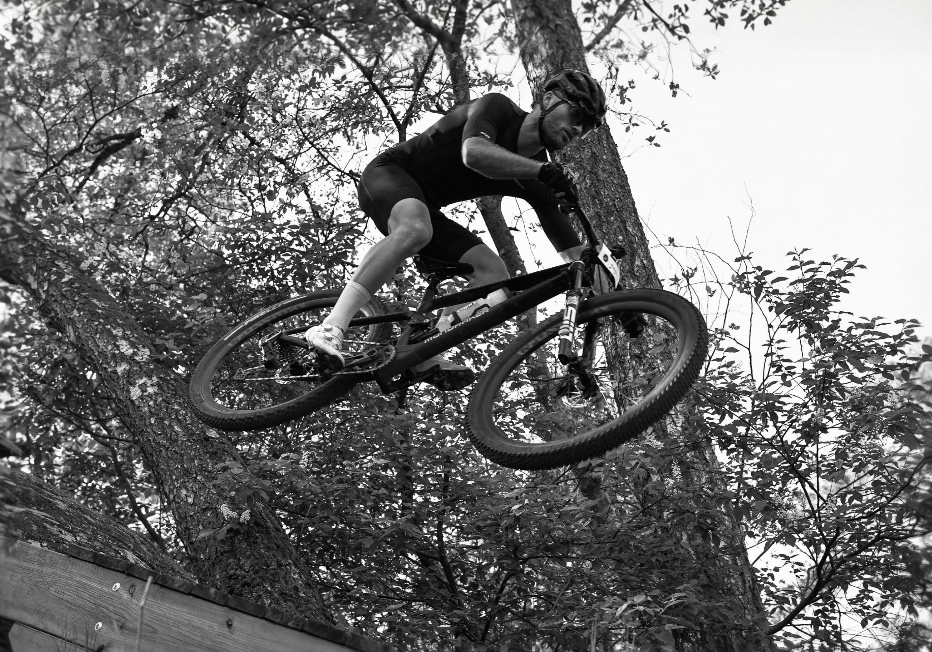 Black and white action shot of a mountain biker mid-jump, leaning over the handlebars while riding a bike in a wooded outdoor setting.