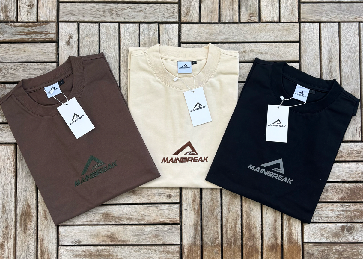 Three folded t-shirts in brown, beige, and black on a wooden surface with 'MAINBREAK' branding.